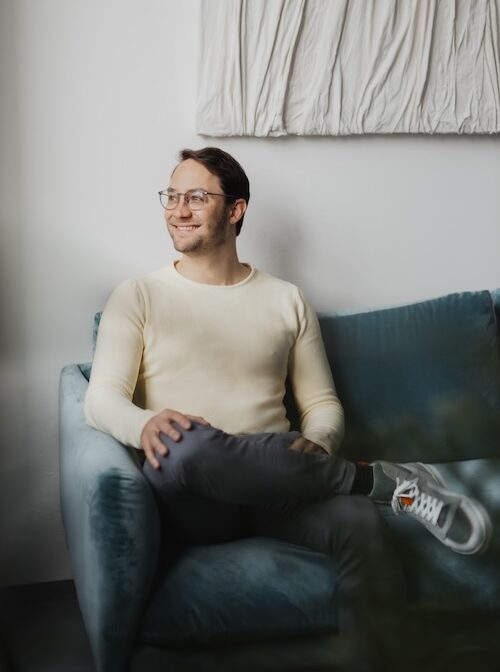 Dr. Thomas Bacharach SIBO Spezialist und Darmgesundheits Experte sitzt auf einem pertolfarbenen Samtsofa in buttergelbem Pullover und mit hellen Sneakers